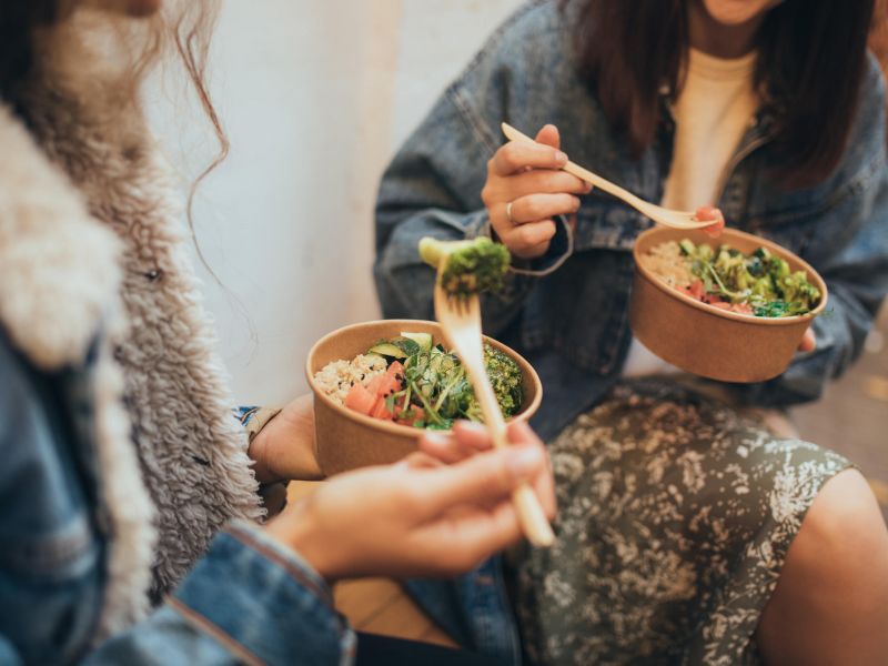 Two people in casual clothes eating salads from bowls with wooden forks while sitting together.