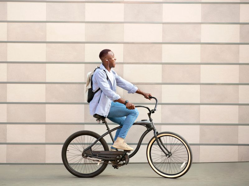 Smiling person with a backpack rides a bicycle past a beige tiled wall, looking ahead.