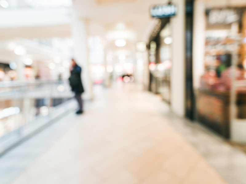 A blurry view of a shopping mall corridor with storefronts and a person standing by a railing.