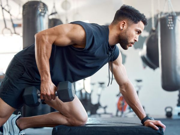 Man exercising with a dumbbell in a gym, doing a one-arm row on a bench with punching bags in the background.