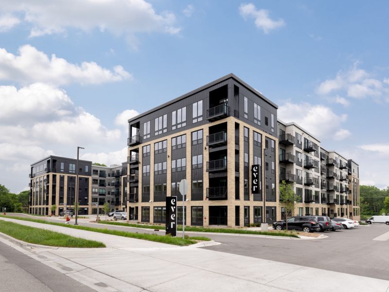 Modern apartment complex with black and beige exterior, balconies, and a parking lot under a partly cloudy sky.