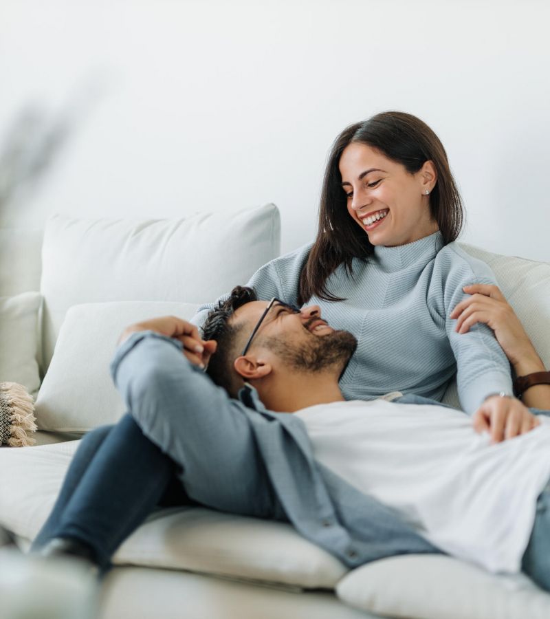 A smiling couple relaxes together on a white couch in a bright living room.