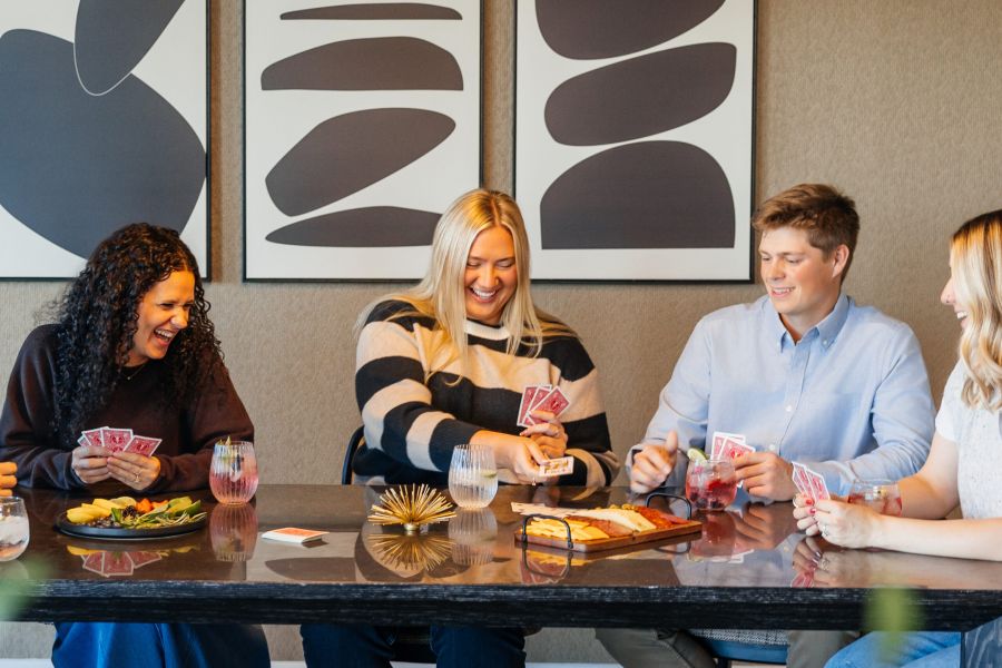 Four people sit at a table, smiling and playing cards, with drinks and snacks in front of them.