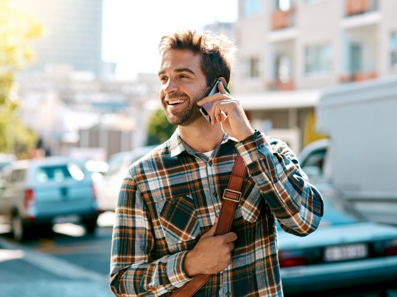 Smiling man in a plaid shirt talking on his phone while walking outdoors on a sunny day.